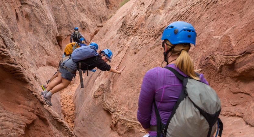 People wearing helmets brace themselves on either side of canyon walls while others look on.